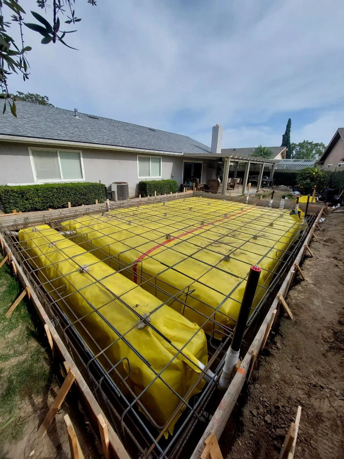 A reinforcing steel grid tied over a yellow vapor barrier inside timber formwork, ready for a slab pour behind a single-story house