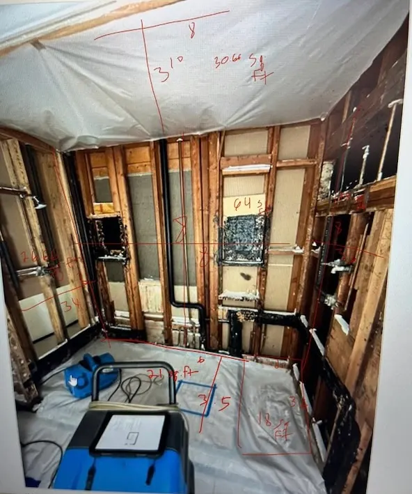 A bathroom stripped back to the studs after a water loss, plastic sheeting over the ceiling and floor, a drying fan running on the floor, and the room’s dimensions written across the photograph in red