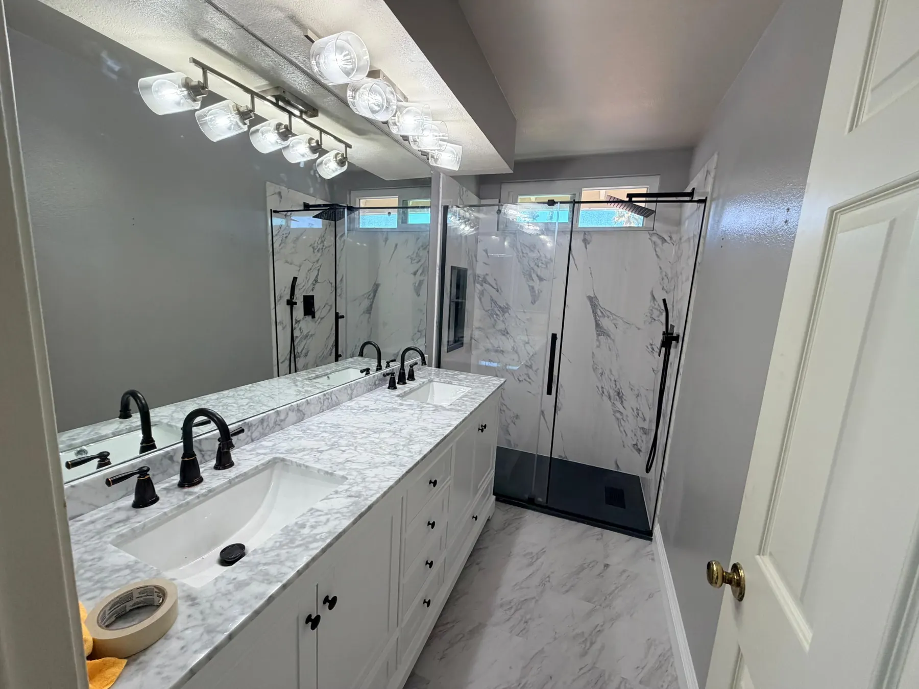 A finished bathroom with a white double vanity in veined stone, black faucets and handles, and a black-framed sliding glass shower against marble-look panels