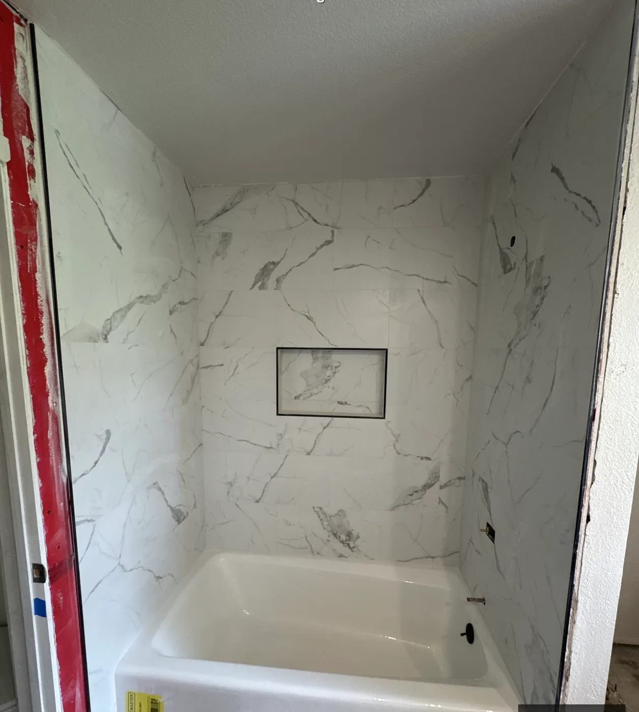 A shower with the marble-look tile set and grouted and the niche trimmed, protective covering still on the tub