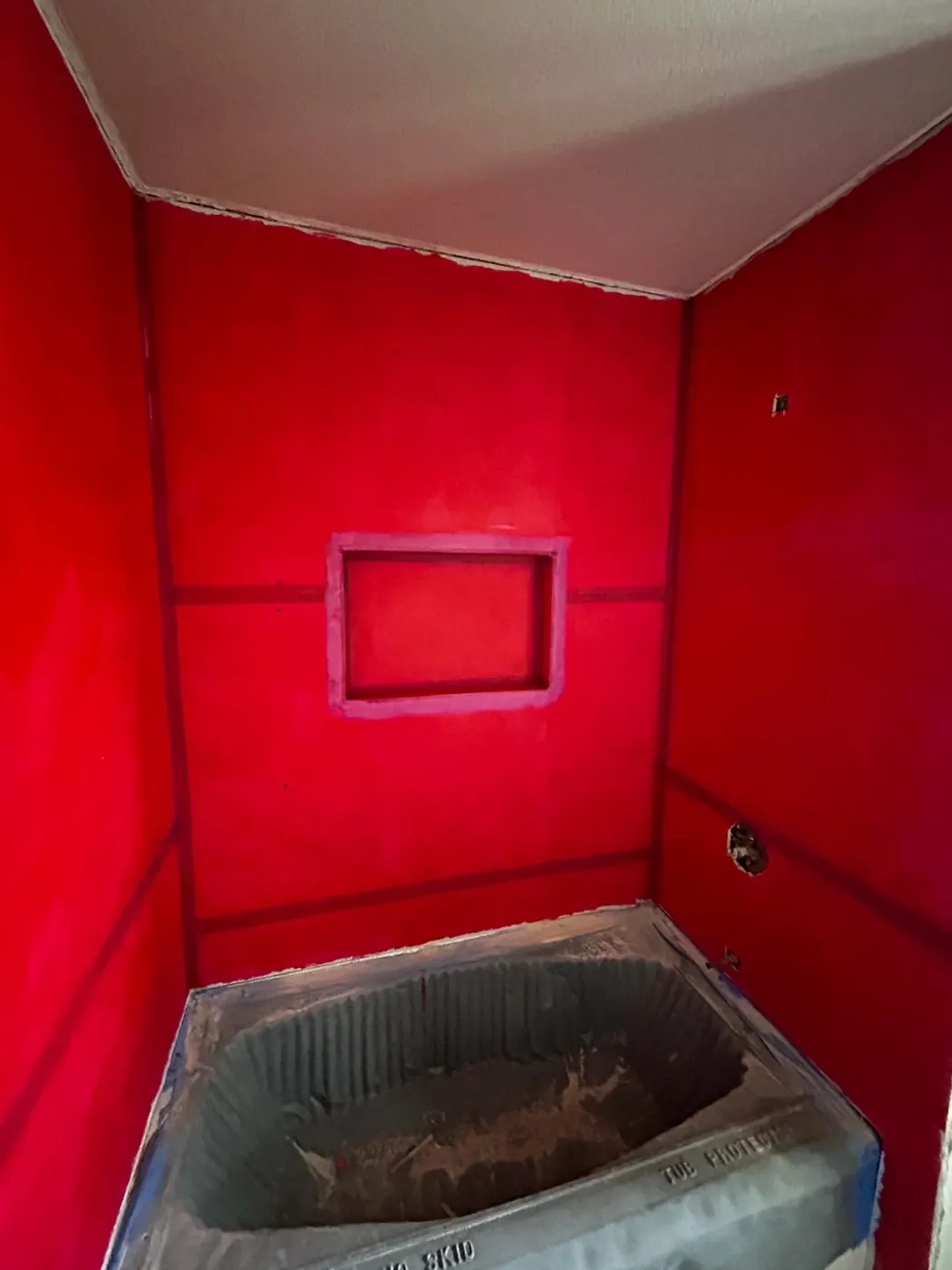 A shower under construction with the walls coated in red waterproofing membrane, the niche formed, and a new tub set and covered below